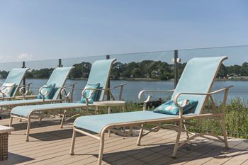 A row of blue lounge chairs with cushions are on a wooden deck by the water.